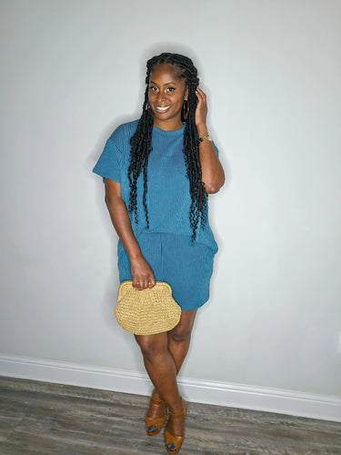 Woman in a blue outfit holding a straw bag against a plain background